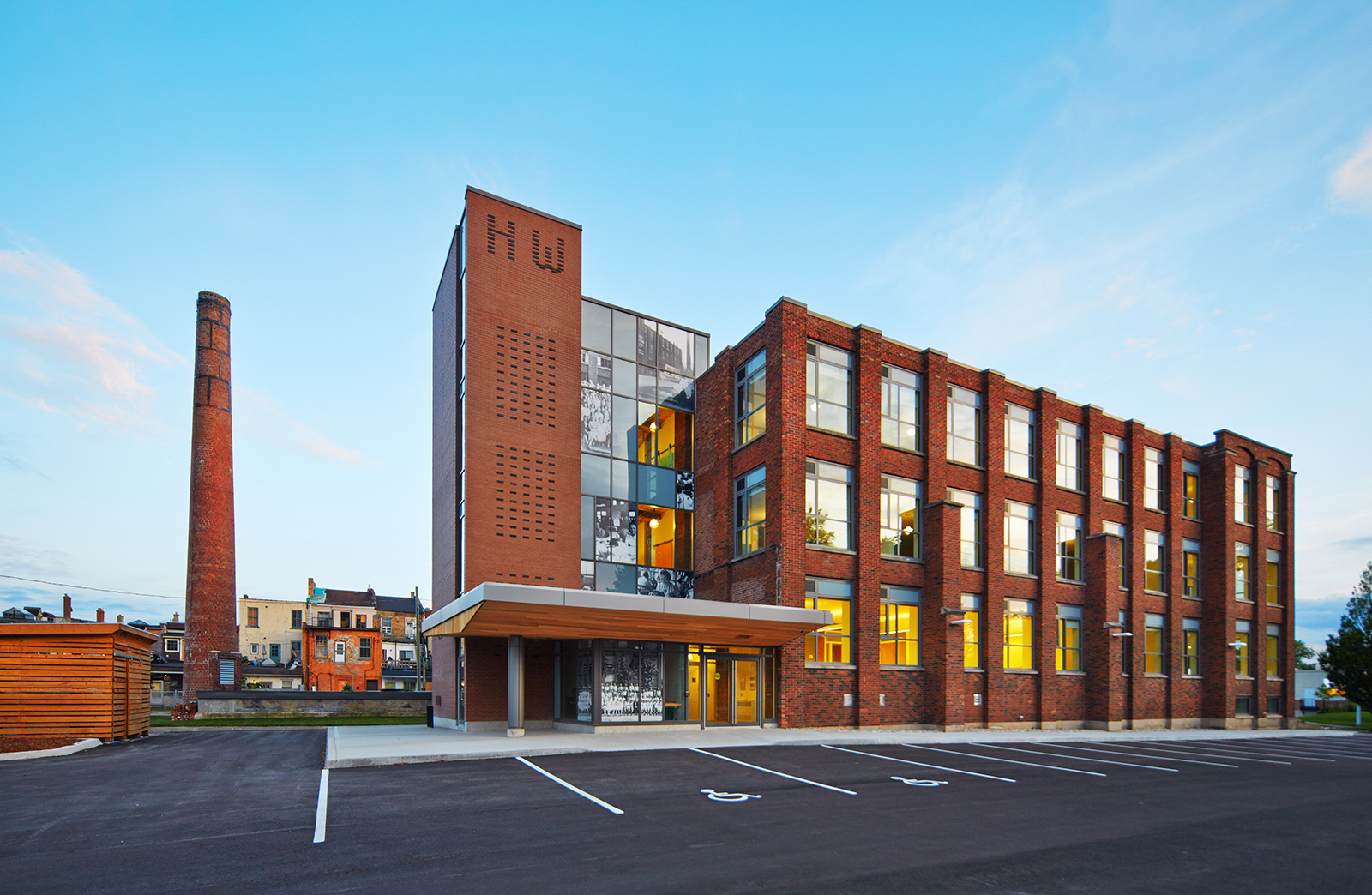 Exterior view of a renovated brick building with large windows and a modern entrance canopy. The building features a vertical brick tower with dark bricks forming the initials ‘HW’ and a glass section with historical photos. A tall, weathered brick smokestack is visible to the left, hinting at the building’s industrial past.